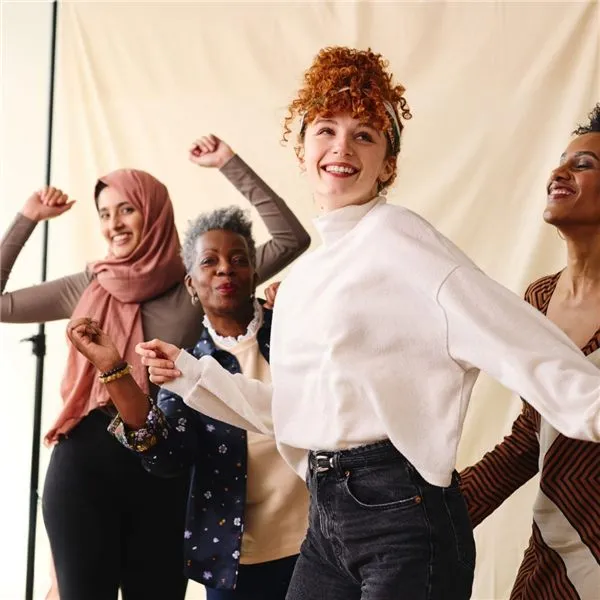 Four women of different ages and backgrounds smiling and dancing together